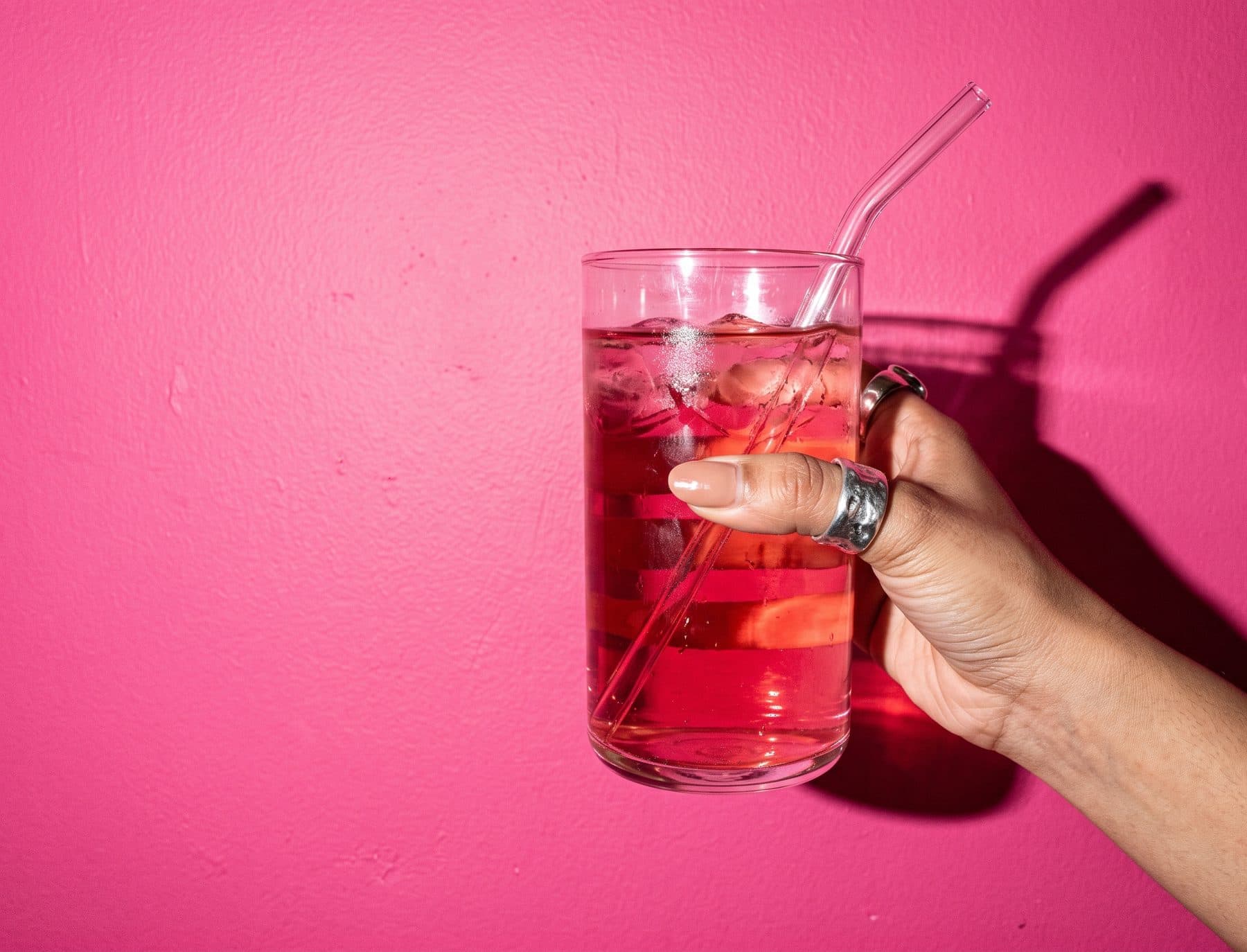 A hand with silver rings holds up a glass of pink water with a glass straw against a hot-pink wall